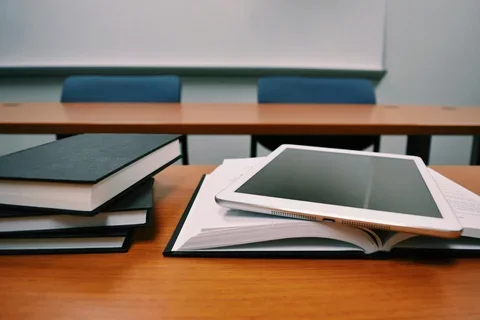 classroom with two long desks. the closest desk is right in frame of the photo and has 3 closed books on the left and one open book with an iPad or some kind of tablet on the right